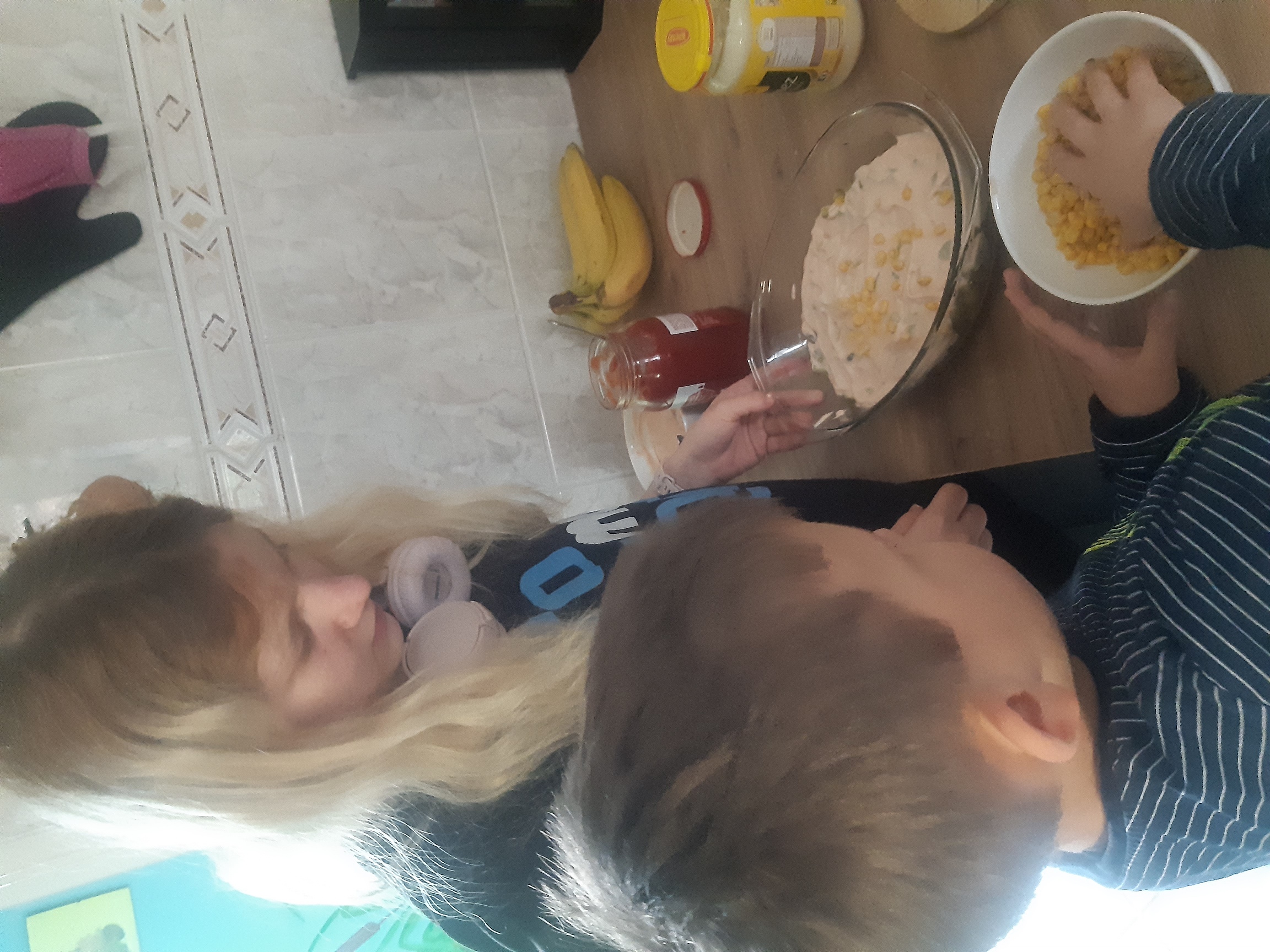 Dwoje wychowanków stoi przy stole, Natan dorzuca kukurydzę do miski z sałatką, którą trzyma Asia