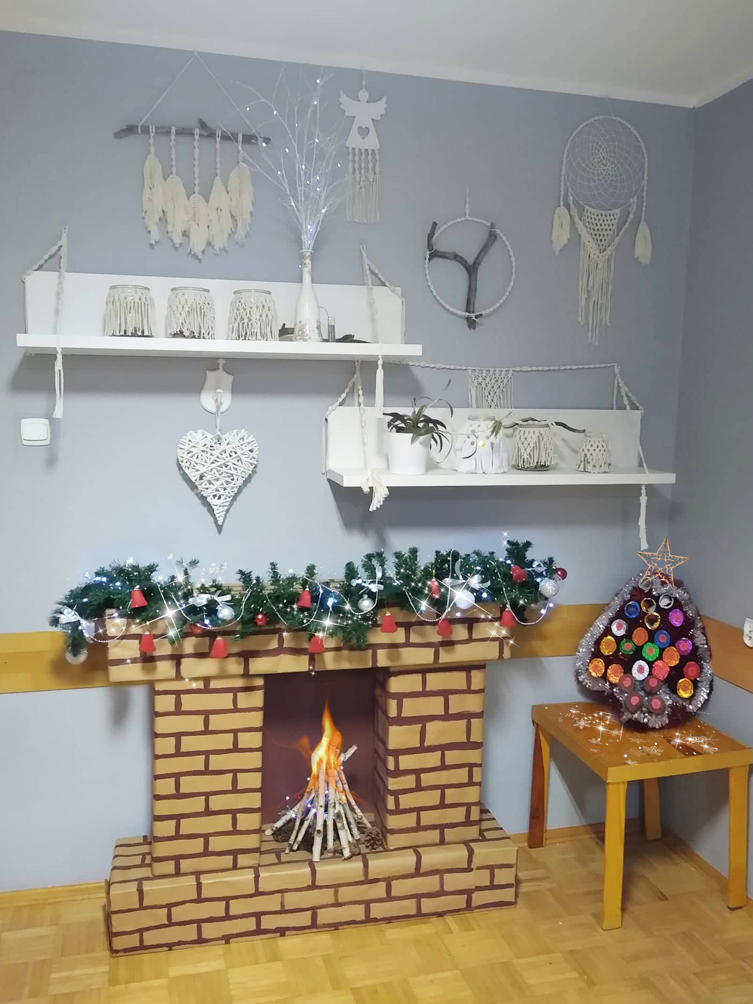 kartonowy kominek udekorowany świąteczną girlandą na tle ściany z ozobami z makramy