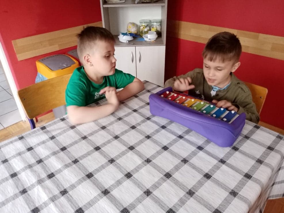 dwaj chłopcy siedzą przy stole, jeden gra na kolorowych metalowych organkach